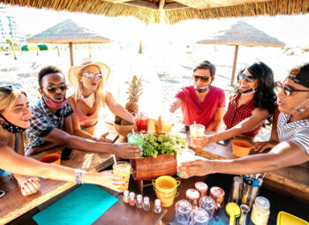 Young trendy people toasting at beach cocktail bar chiringuito with open mask - New normal summer life style concept with friends having fun together cheering drinks - Warm bright backlight filter